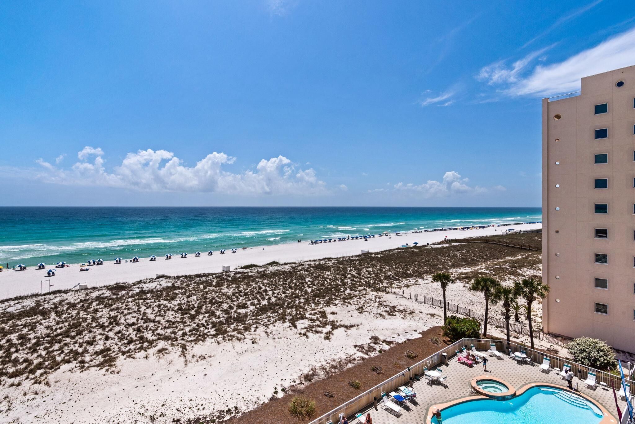 A beach view with a pool area, a building with multiple floors, and a sandy beach.