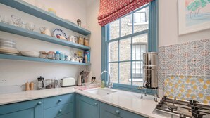 Una cocina con armarios azules, encimera blanca, ventana con un valance rojo y azulejos de salpicadero con estampado.