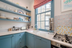 A kitchen with blue cabinets, a white countertop, a window with a red valance, and patterned backsplash tiles. - Gorgeous 3 bedroom flat - Pimlico (Greater London)
