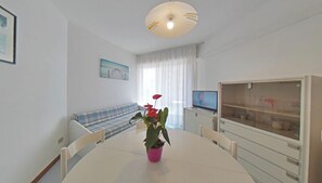 A dining area with a round table, chairs, a potted plant, a television, and a sofa. - Sea View Stay in a Beachfront Building With Pools (Lignano Sabbiadoro)