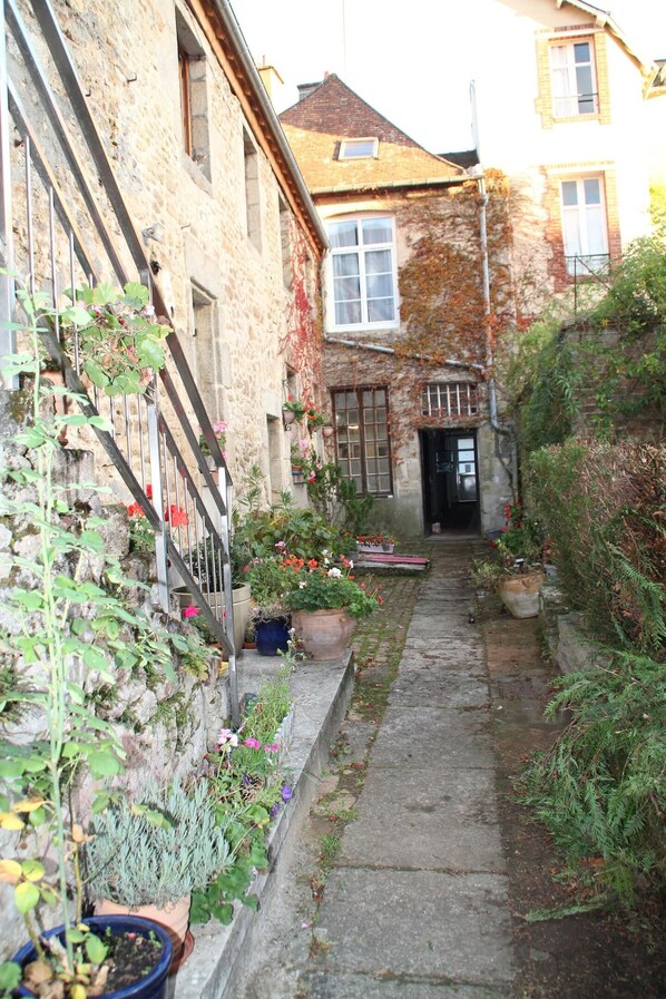 Un étroit sentier de pierre mène à un bâtiment doté d’une porte et de fenêtres en bois, qui est entouré de plantes et de fleurs en pot.