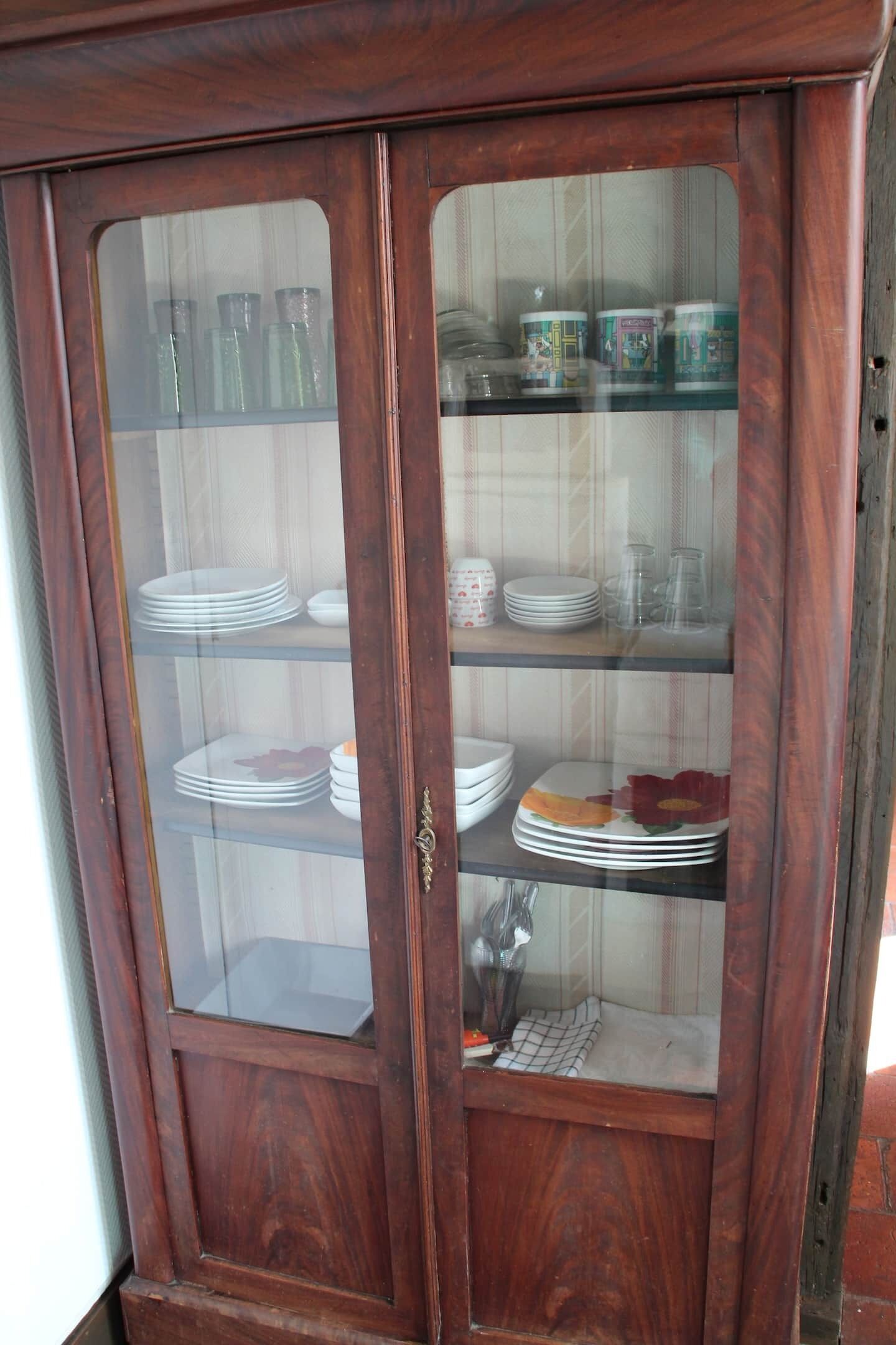 A wooden cabinet with glass doors, containing stacked plates and glassware.