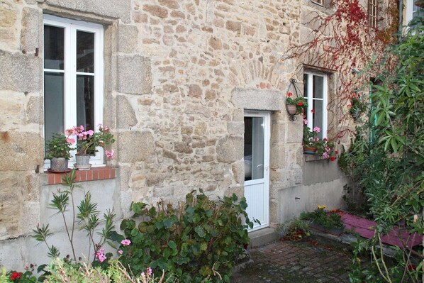 A stone building with two windows, one with a door, and potted plants on the windowsills.