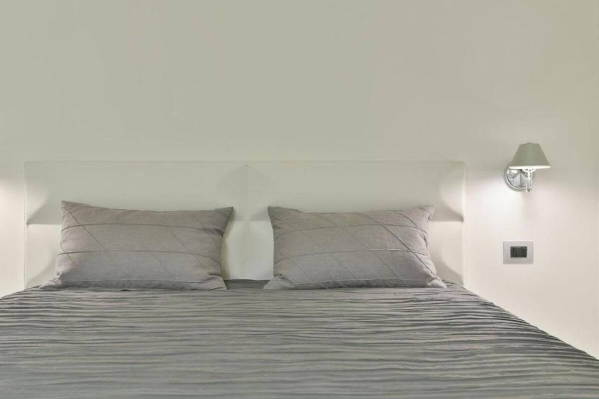 Un lit avec une tête de lit, deux oreillers, une lampe fixée au mur et un petit interrupteur sur le mur.