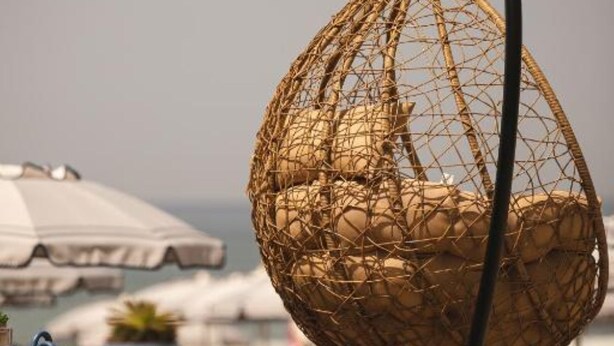 A wicker hanging chair with a chain, a potted plant, and a beach umbrella on a wooden deck. - Royal Rooms (Laura)