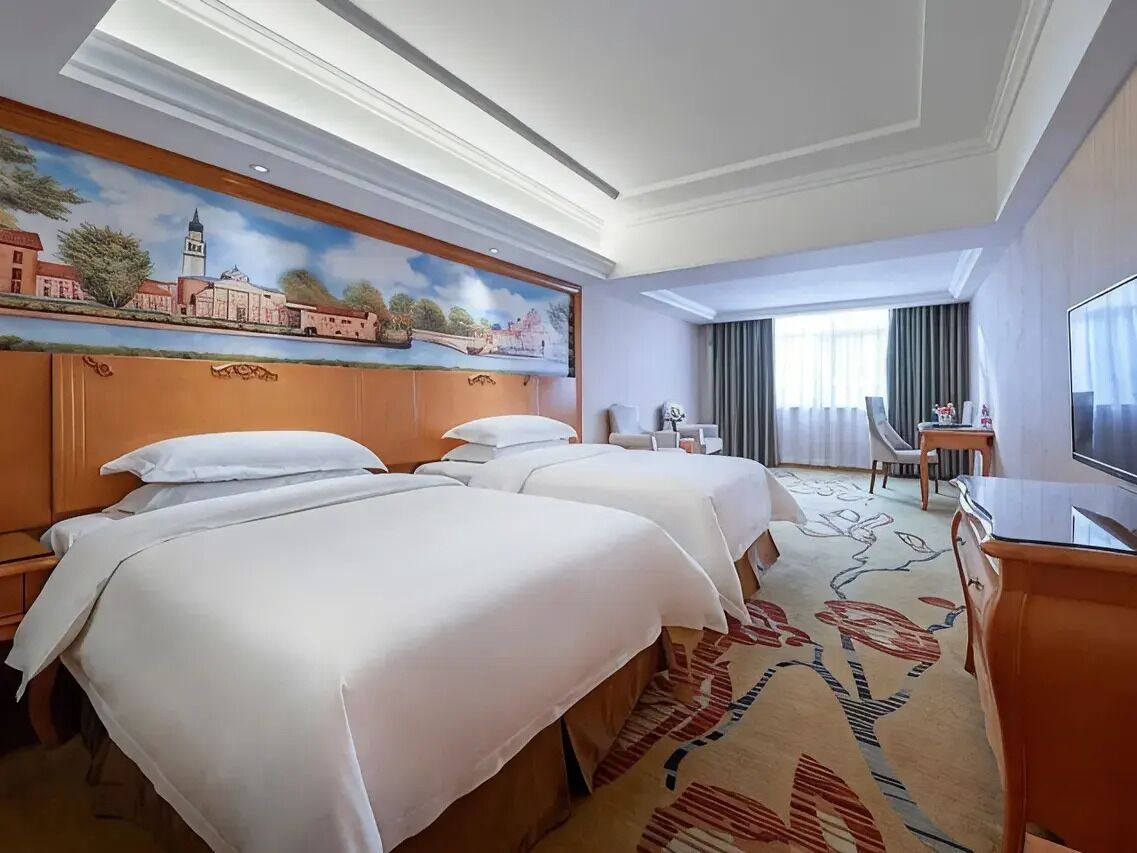 Ein Hotelzimmer mit zwei Betten, einem Fernseher, einem Schreibtisch und einem Bild an der Wand.