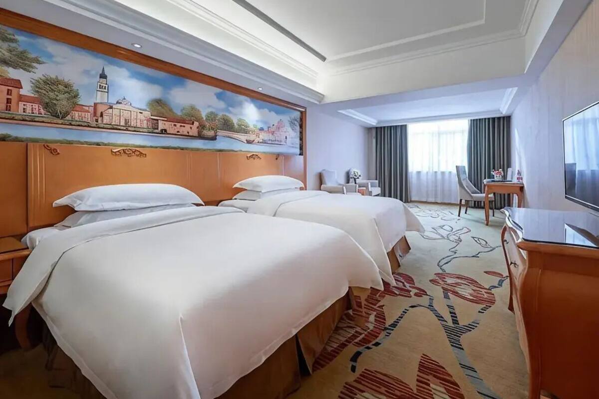 A hotel room with two beds, a television, a desk, and a painting on the wall.