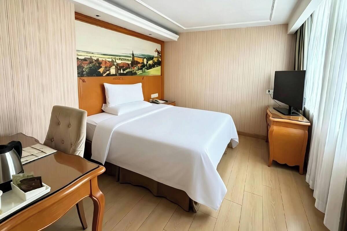 A hotel room with a bed, a desk with a kettle, a television, and a painting on the wall.