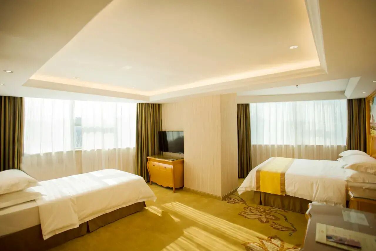 Ein Hotelzimmer mit zwei Betten, einem Fernseher, einem Schreibtisch und einem großen Fenster mit Vorhängen.