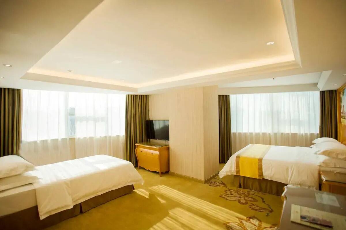 A hotel room with two beds, a TV, a desk, and a large window with curtains.