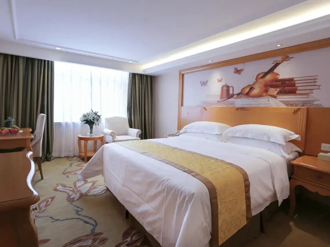 Ein Hotelzimmer mit Bett, Sessel, kleinem Tisch mit Vase und einem Wandbild.
