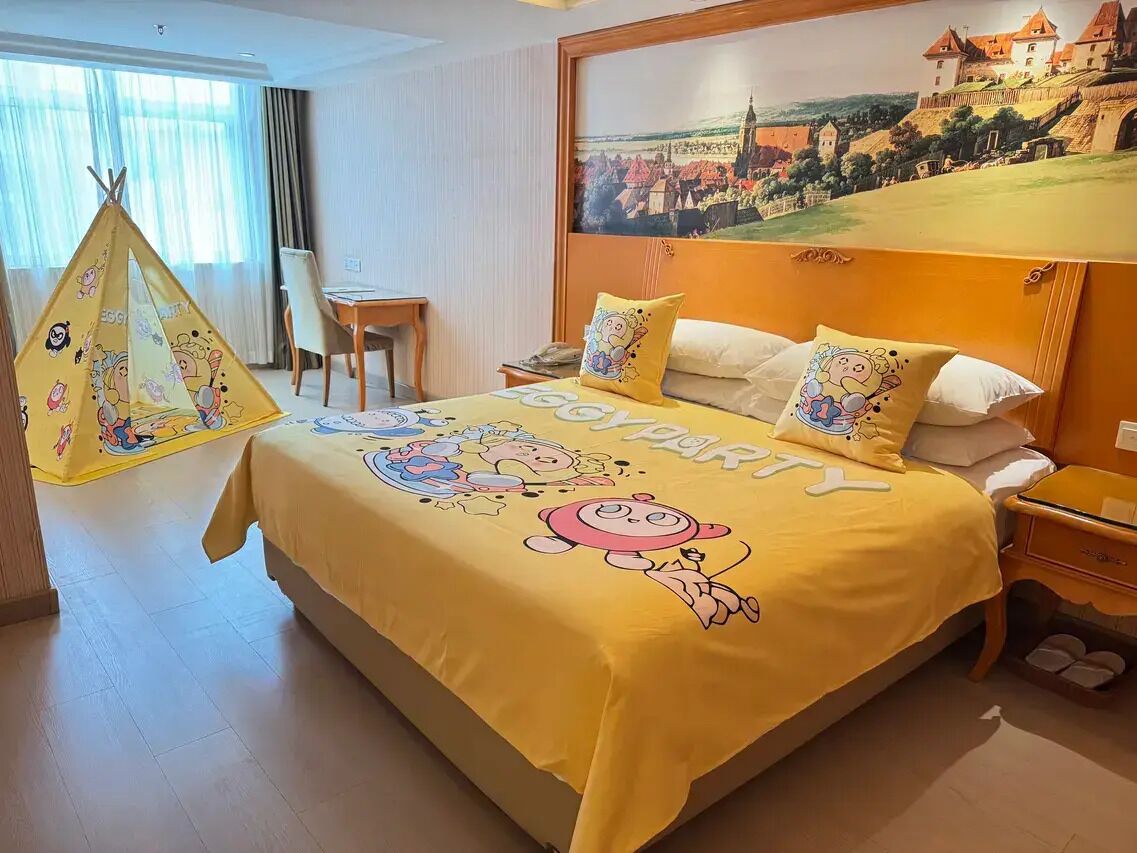 Ein Hotelzimmer mit einem Bett, das mit einem 'Jungenparty'-Themenüberwurf bezogen ist, einem Essbereich mit Tisch und Stühlen sowie einem großen Fenster mit Blick auf eine Burg.