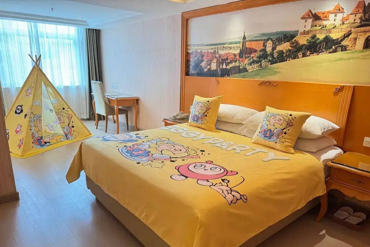 A hotel room with a bed featuring a 'boy party' themed bedspread, a dining area with a table and chairs, and a large window with a view of a castle.