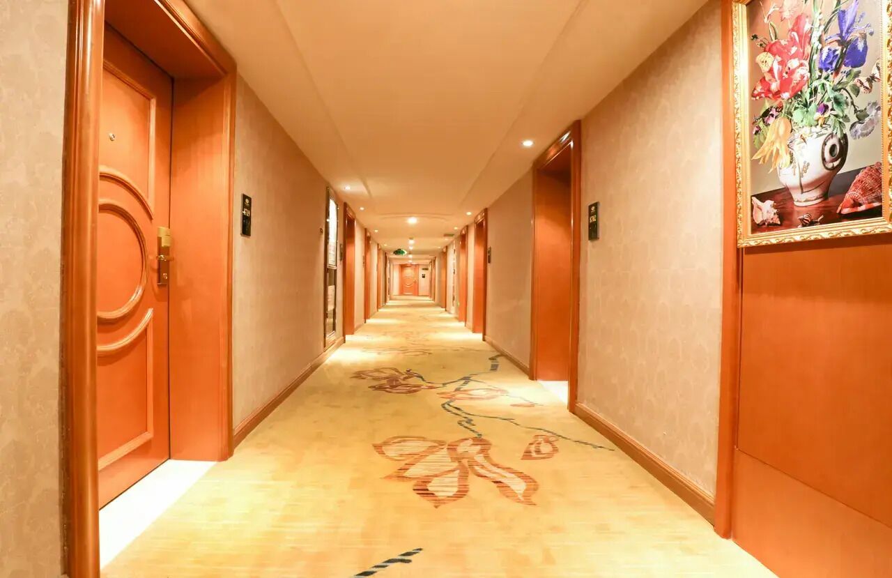 A hotel corridor with wooden doors, a carpeted floor, and a floral painting on the wall.