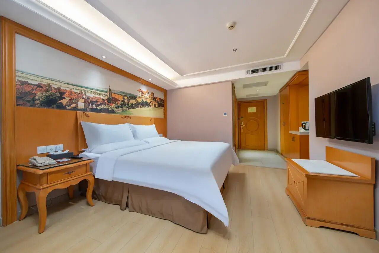 A hotel room with a bed, a TV, a desk, a nightstand, and a painting on the wall.