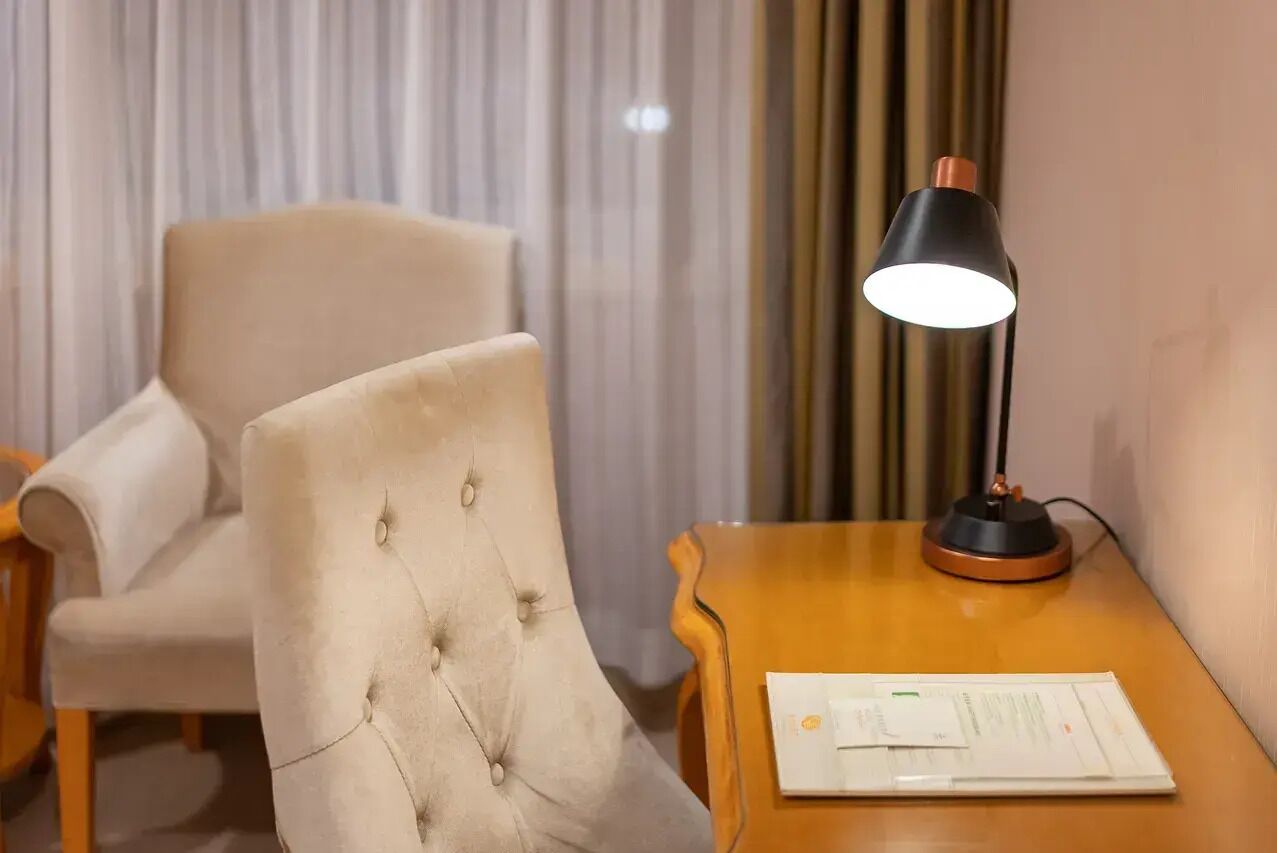 A room with a tufted armchair, a wooden side table with a lamp, and a window with curtains.