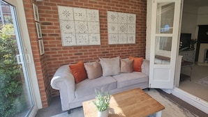 Una habitación con pared de ladrillo, un sofá blanco con cojines anaranjados, una mesa de centro de madera y una puerta que da a otra habitación.
