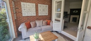 A room with a brick wall, a white sofa with orange cushions, a wooden coffee table, and a door leading to another room. - ✨Parc Avenue – A Charming Stay in the Heart of Cosham, Portsmouth! ✨ (Cosham)