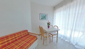A room with a sofa, a dining table with chairs, a potted plant, and a framed picture on the wall. - Ideally Located sea Front Building With Pool (Lignano Sabbiadoro)