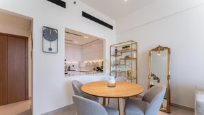 A modern dining area with a round wooden table, four grey chairs, a wall-mounted clock, a gold-framed mirror, and a kitchen visible through an open doorway. - Layover Holiday Homes (Dubai)