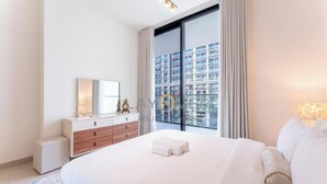 A modern hotel room with a large bed, a dresser, a mirror, and a view of a cityscape through a large window. - Layover Holiday Homes (Dubai)