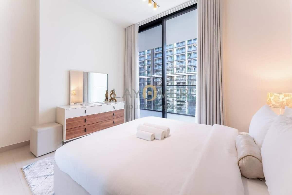 Una habitación de hotel moderna con una cama grande, un aparador, un espejo y vistas a un paisaje urbano a través de un ventanal.
