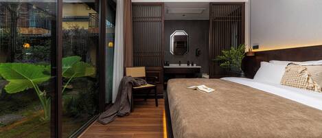 A modern hotel room with a large bed, a desk, a mirror, and a view of a garden through a glass door.