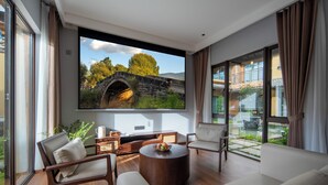 A modern living room with a large TV screen, comfortable seating, and a view of a stone bridge and greenery.