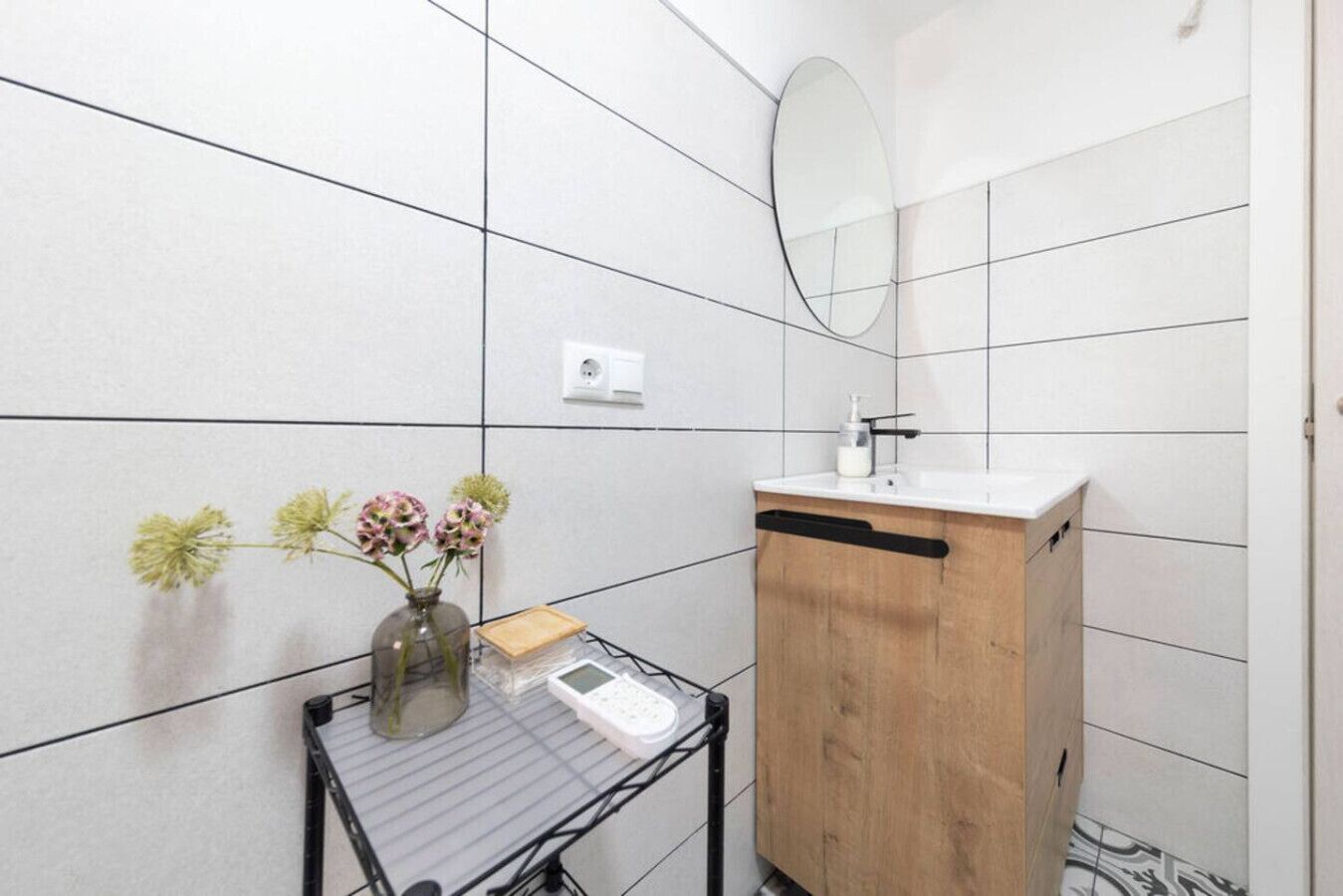 Un baño con paredes de azulejos blancos, un mueble de madera, un lavabo, un espejo, una mesita con un jarrón de flores y una balanza.