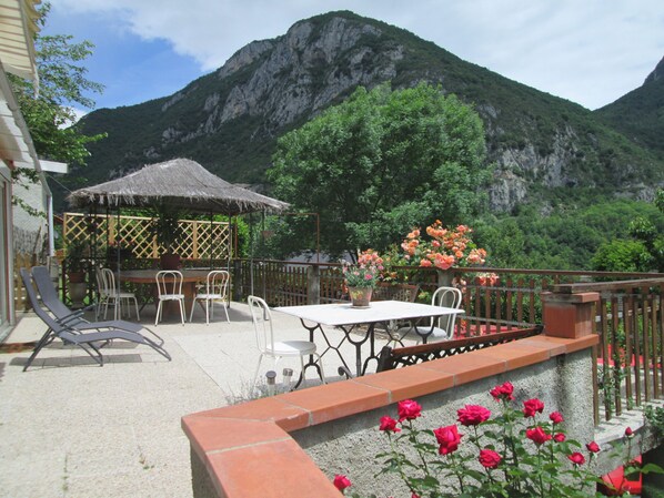 Une terrasse extérieure avec une table et des chaises, une structure dotée d’un toit de chaume et une vue sur les montagnes en arrière-plan.