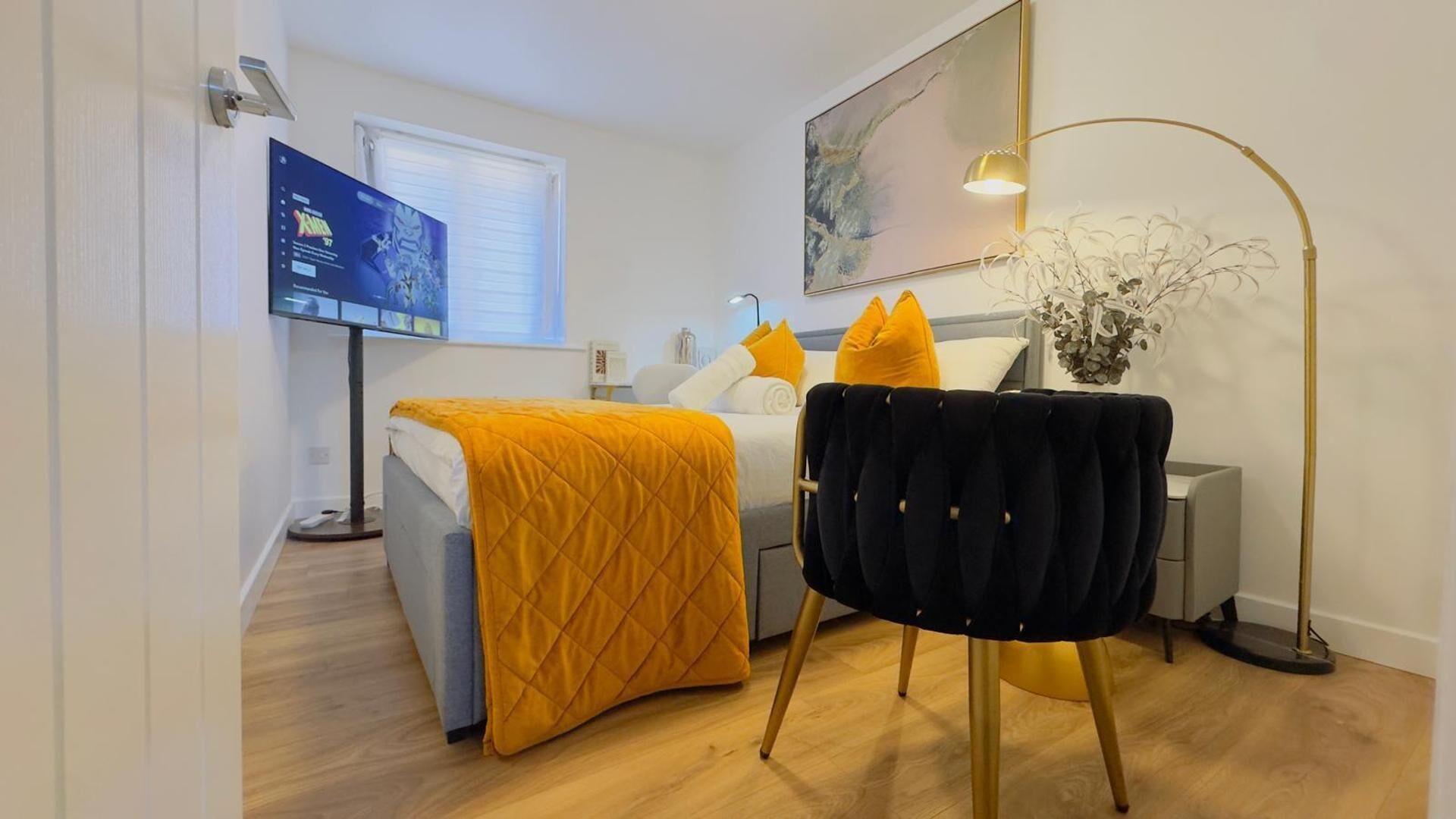 A modern hotel room with a bed, a desk with a computer, a chair, a floor lamp, and a painting on the wall.