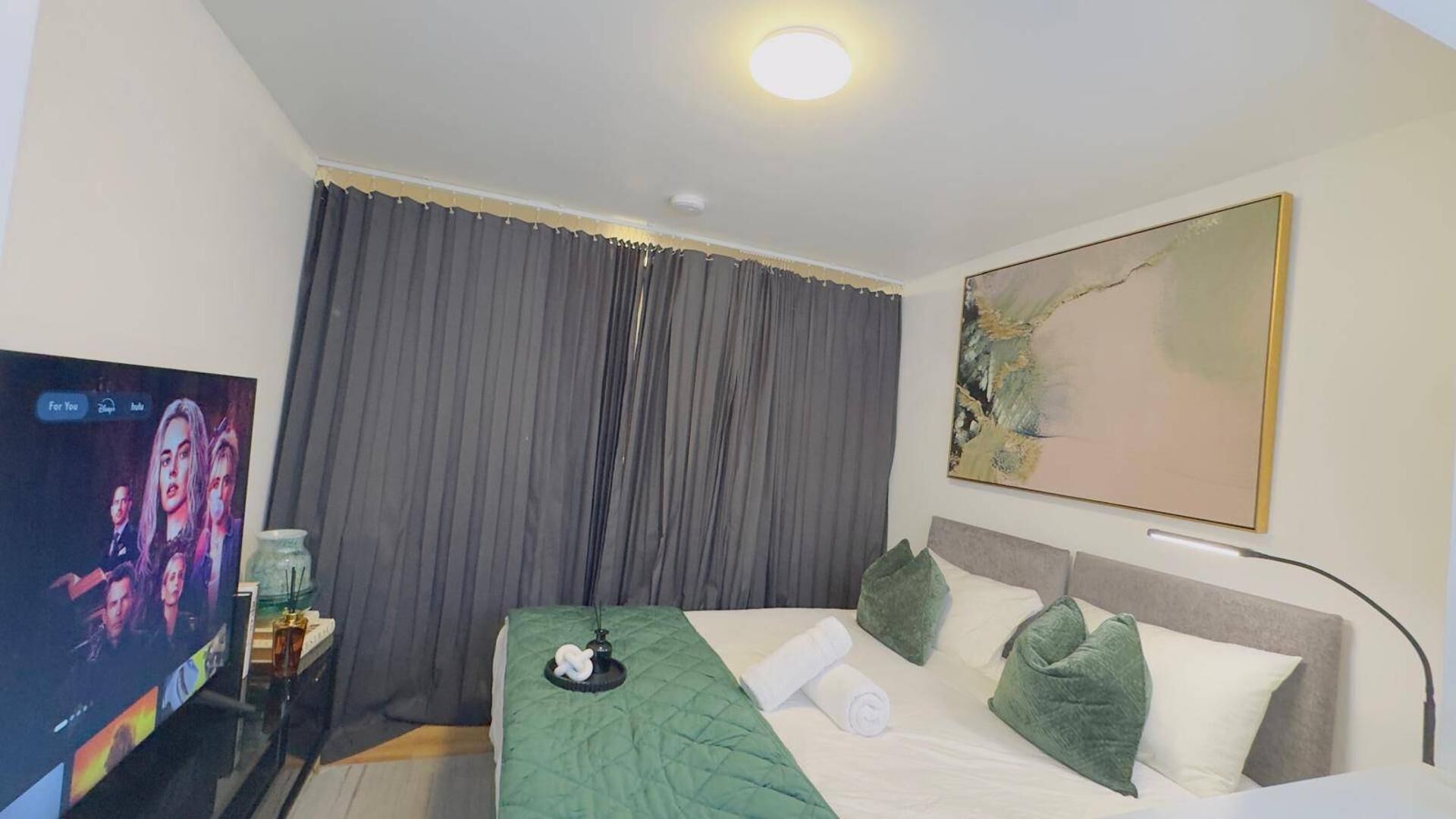 A hotel room with a bed, a TV, a painting, and a window with curtains.