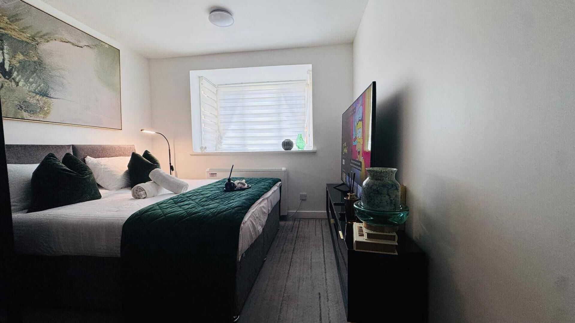 Une chambre à coucher avec un lit, une fenêtre avec des stores, une lampe, un cadre photo et un vase posé sur une table de chevet.