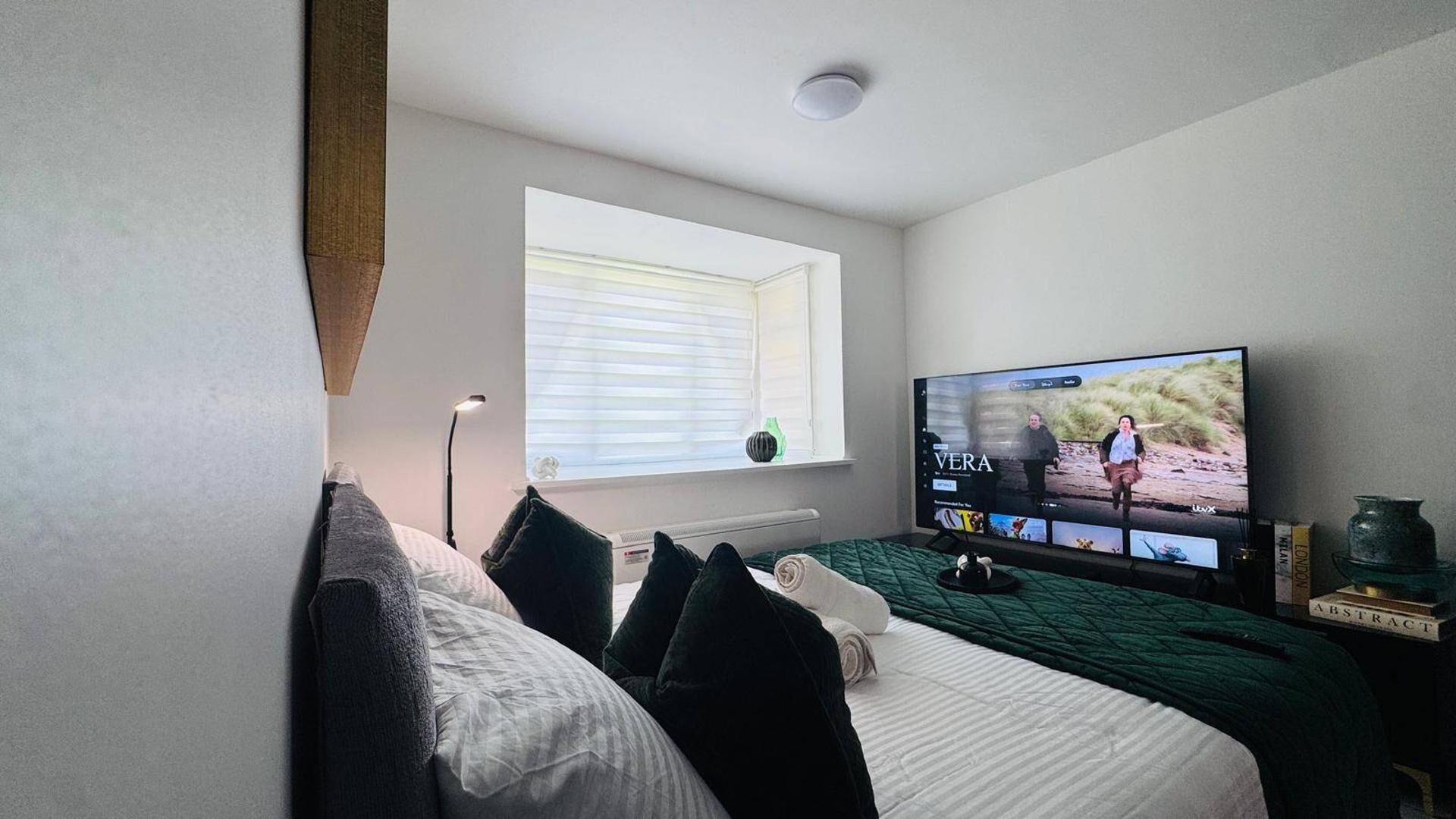 A bedroom with a bed, a window, a television, and a bedside table with books.