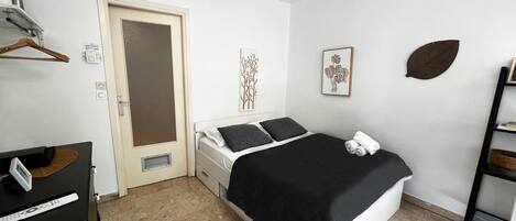 A bedroom with a bed, a desk, a chair, a wardrobe, a door, and a shelf with a plant.