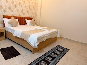 A neatly made bed with a headboard, a bedside table, a dark rug, and a patterned rug on the floor. - The Classic Crown (Ayodhya)