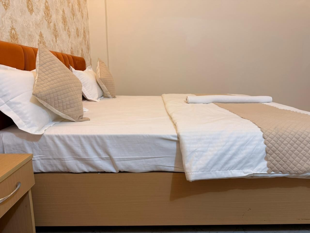 A double bed with white bedding and a quilted pillow, a wooden nightstand, and a patterned wall.