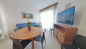 A dining area with a round wooden table, chairs, a potted plant, a blue sofa, a TV, and a painting on the wall. - Family Base on the sea Front With Pool Access (Lignano Sabbiadoro)