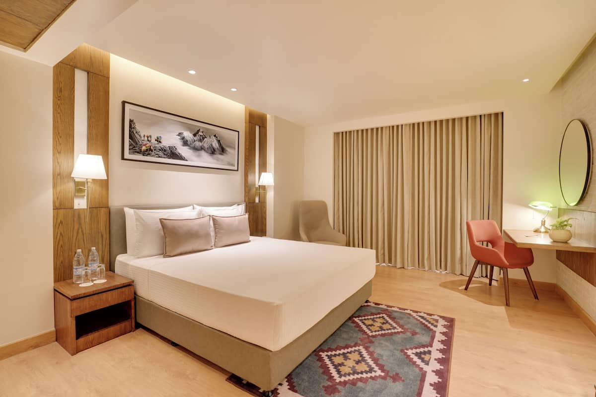 Una habitación de hotel con cama, mesita de noche, escritorio, silla y un ventanal con cortinas.