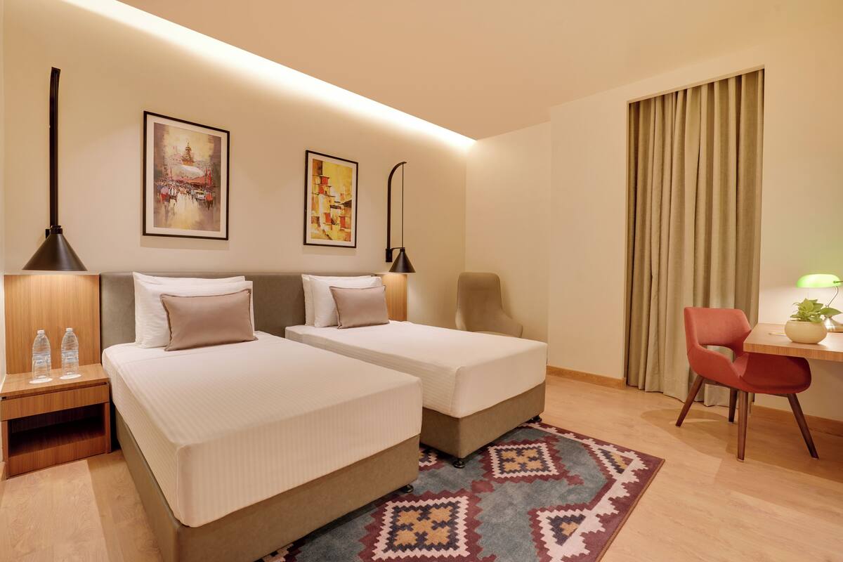 Habitación de hotel con dos camas, un escritorio, una silla, una lámpara y dos cuadros en la pared.