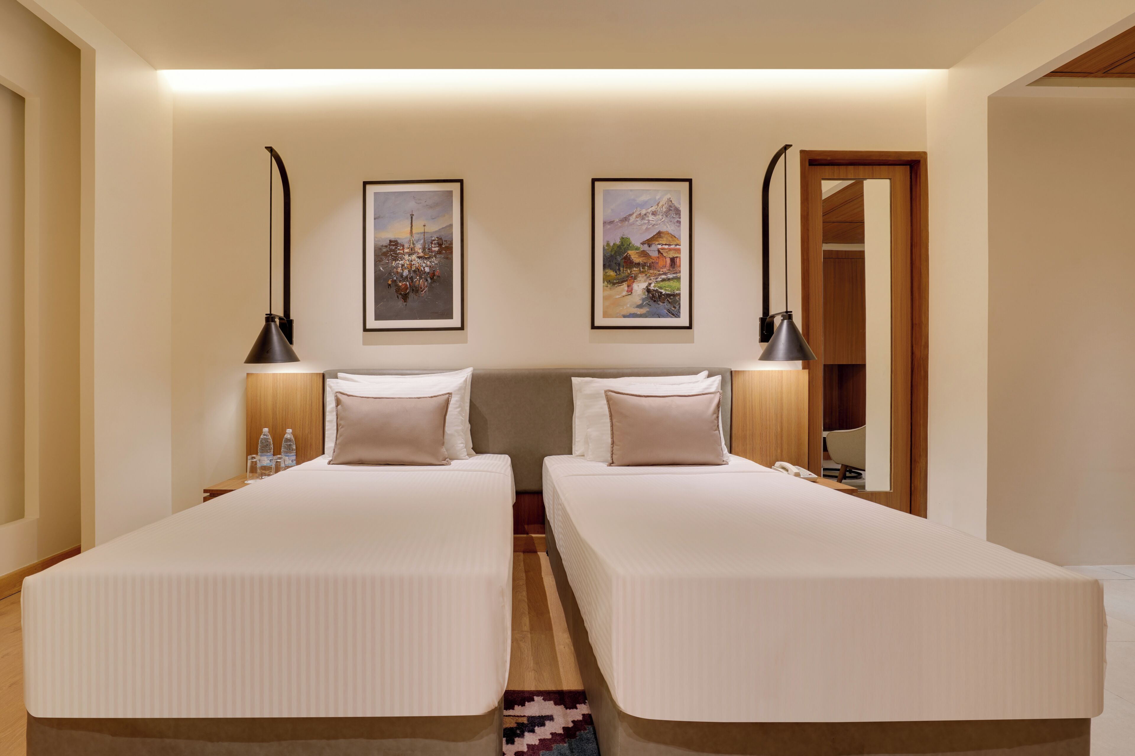 A hotel room with two single beds, a wooden floor, a rug, two wall-mounted lamps, and two framed pictures.