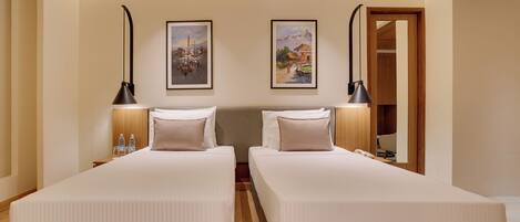 A hotel room with two single beds, a wooden floor, a rug, two wall-mounted lamps, and two framed pictures.