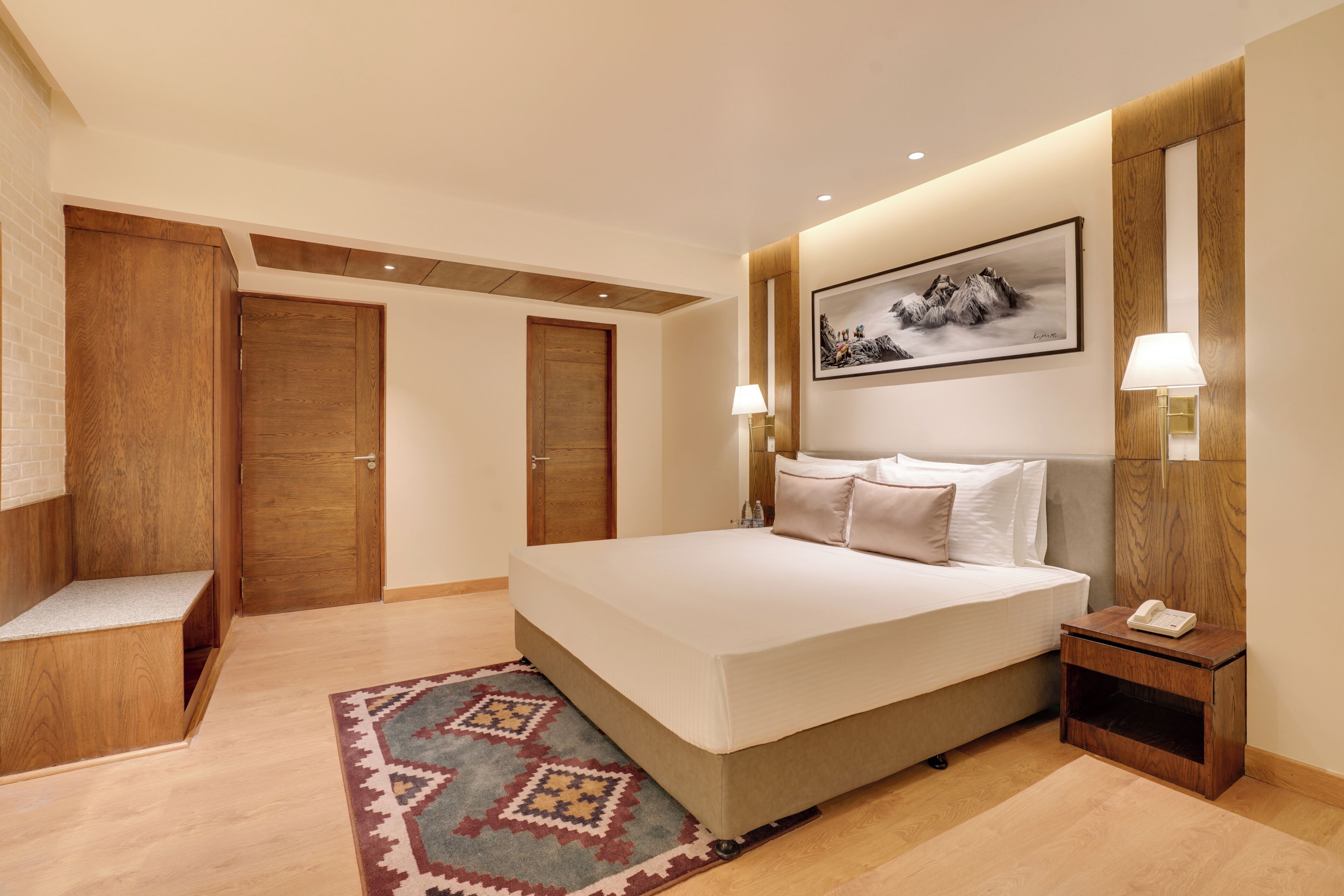 A hotel room with a large bed, a bedside table with a lamp, a wooden bench, and a patterned rug.