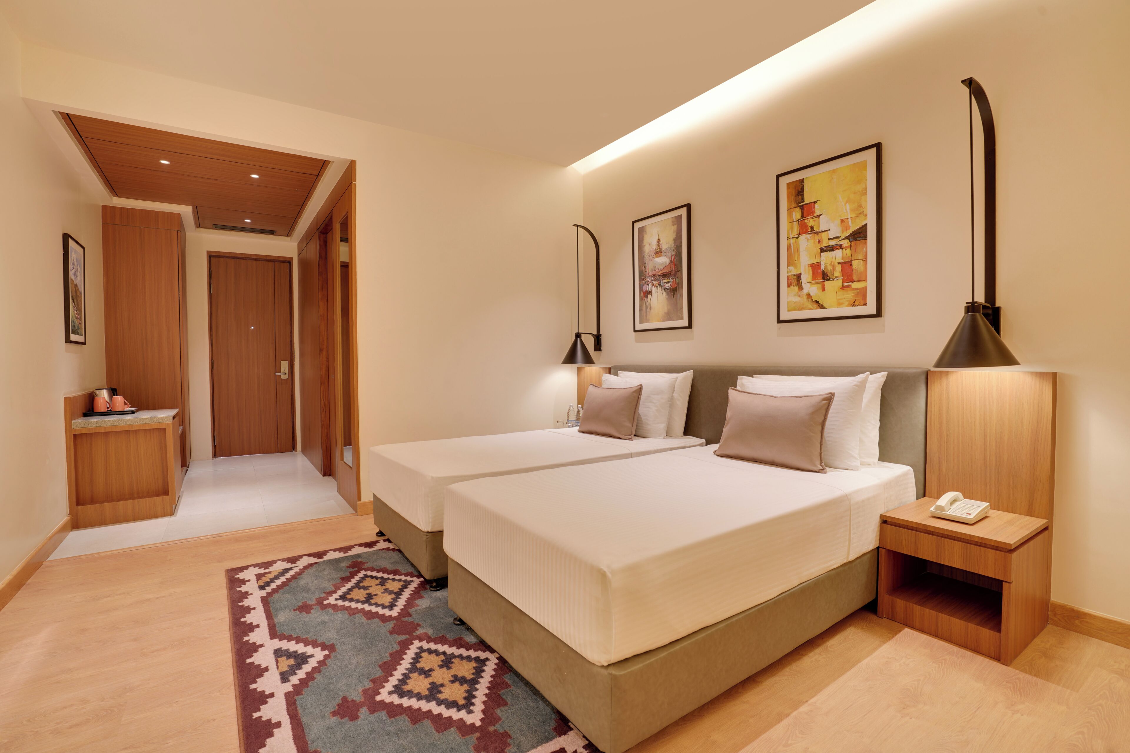 Habitación de hotel con dos camas, una mesita de noche con lámpara, un escritorio pequeño con teléfono y una alfombra estampada.