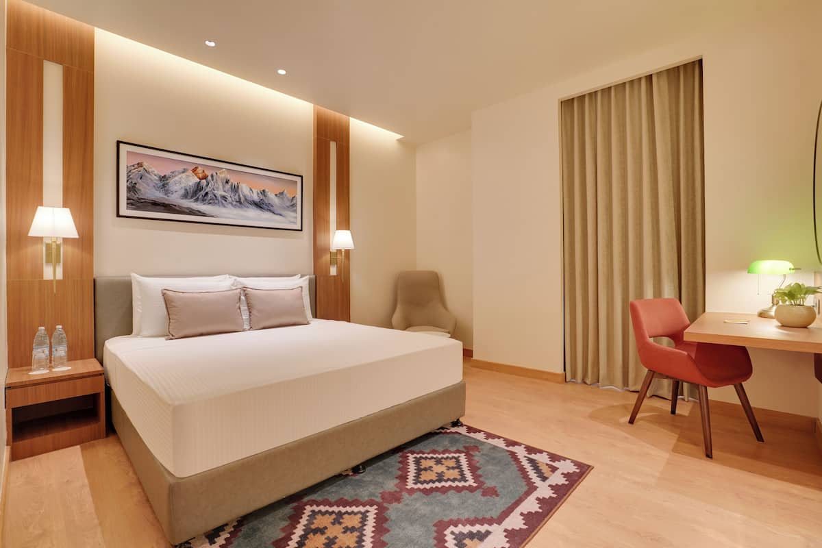 Habitación de hotel con cama, escritorio, silla, lámpara, botella de agua y un cuadro en la pared.