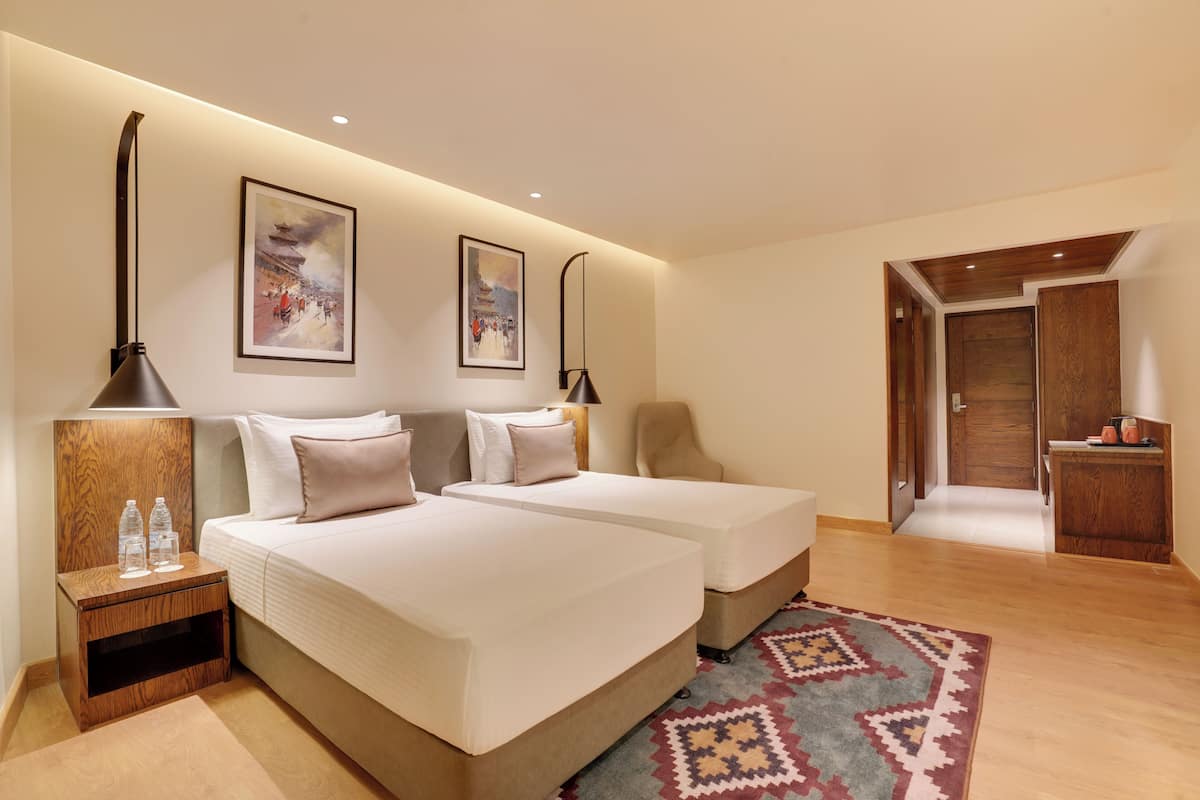 Habitación de hotel con dos camas, una mesita de noche con botellas de agua, una alfombra estampada y obras de arte en las paredes.