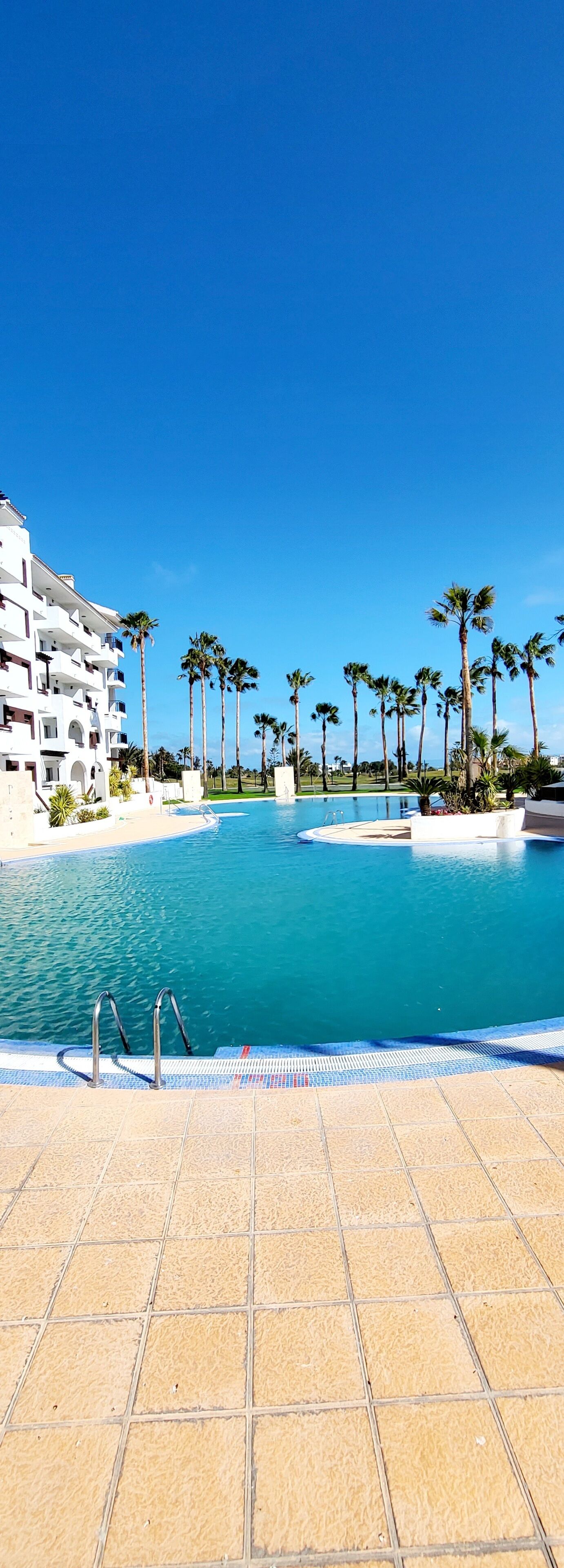 Piscina de un resort con agua azul cristalina, rodeada de palmeras y un edificio blanco con balcones.