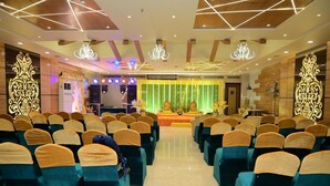 A conference room with rows of chairs, a stage with a backdrop, and decorative lighting fixtures. - Hotel Gurveer Royal (Lucknow)