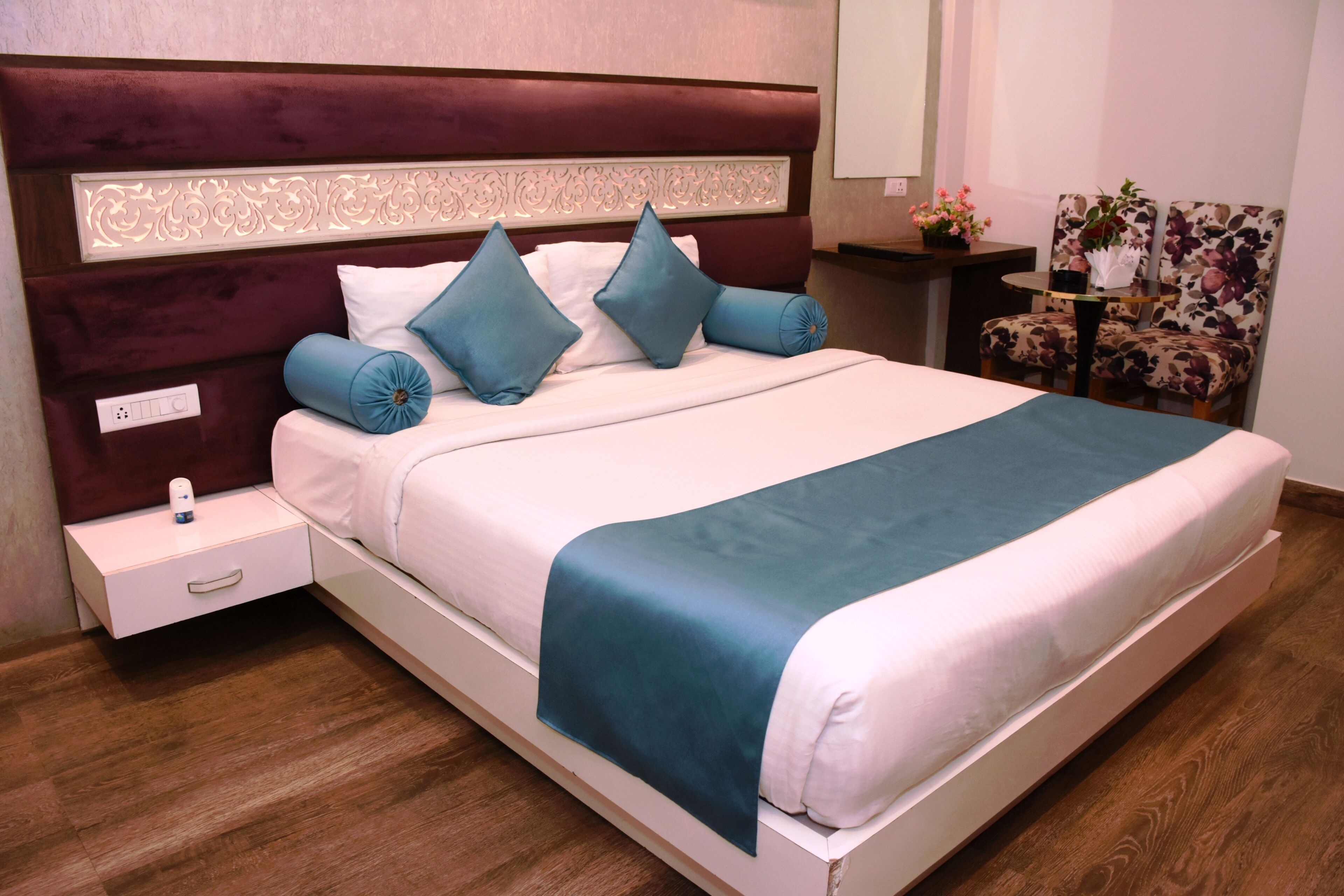 A neatly made bed with a headboard, a bedside table with a lamp, and a floral chair.