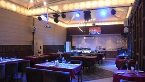 A restaurant with multiple tables, chairs, and a kitchen area. - Hotel Gurveer Royal (Lucknow)